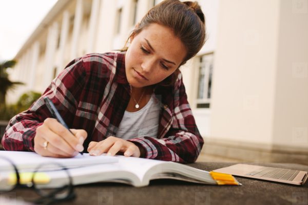 Young woman studying outdoors on a university campus, deeply focused on writing in her notebook next to a laptop and textbooks.